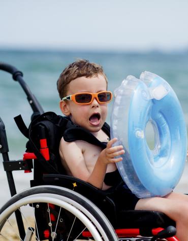 Boy in wheelchair happily holding inflatable tube in a beach setting