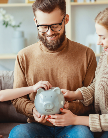 Two adults holding piggy bank while child puts money in it